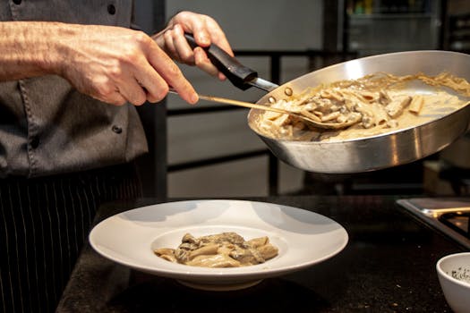 Chef carefully plates a creamy mushroom pasta dish in an indoor kitchen setting.