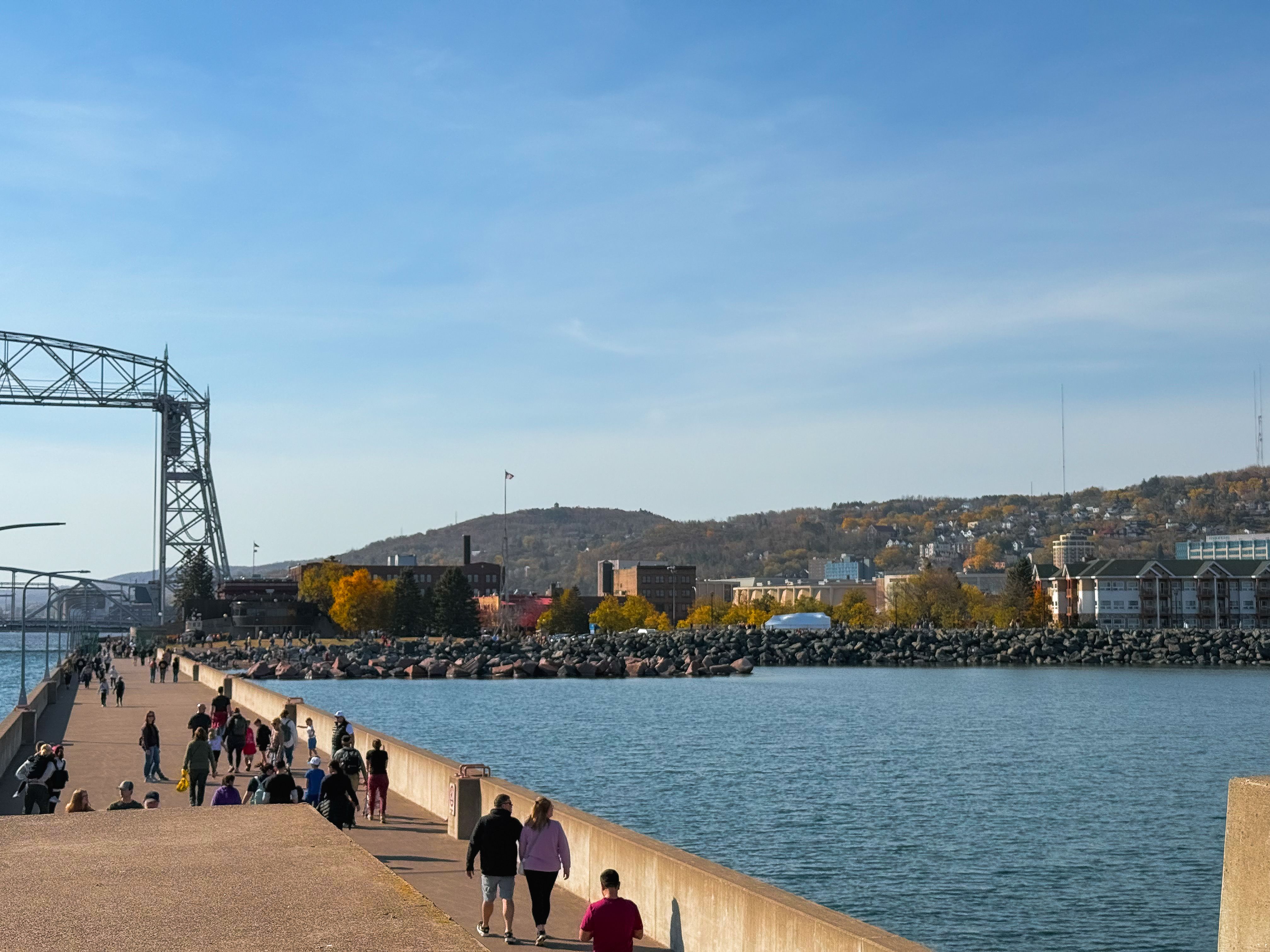 People enjoy a sunny day by the Duluth Aerial Lift Bridge with vibrant fall foliage.