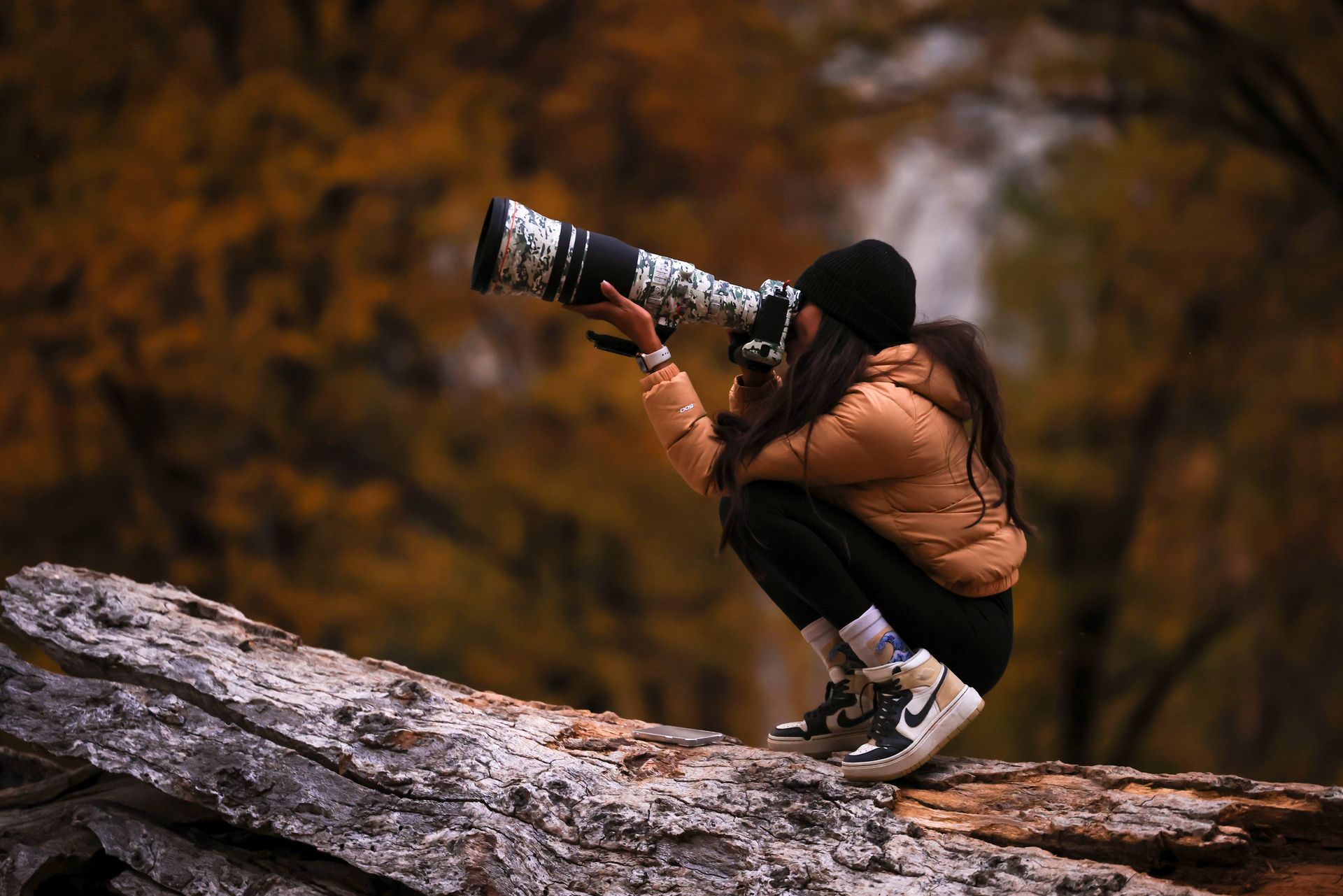 A woman in a brown jacket with a telephoto lens capturing outdoor scenery during fall.