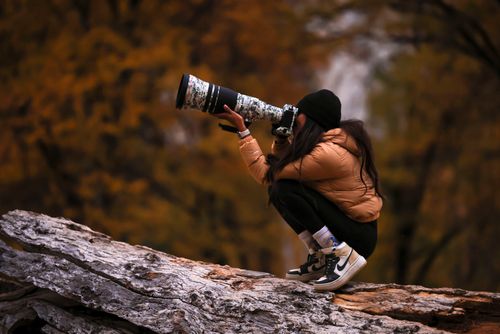 Free A woman in a brown jacket with a telephoto lens capturing outdoor scenery during fall. Stock Photo