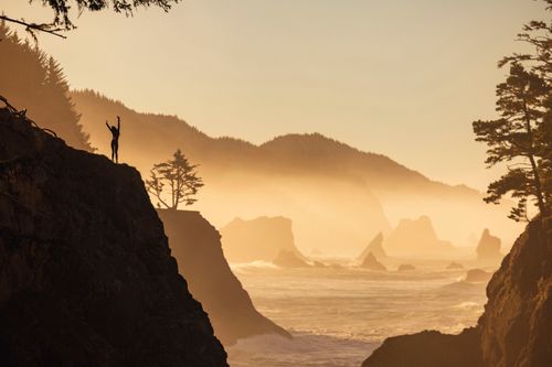 Free A person stands triumphantly on a coastal cliff, enjoying a breathtaking sunrise view. Stock Photo