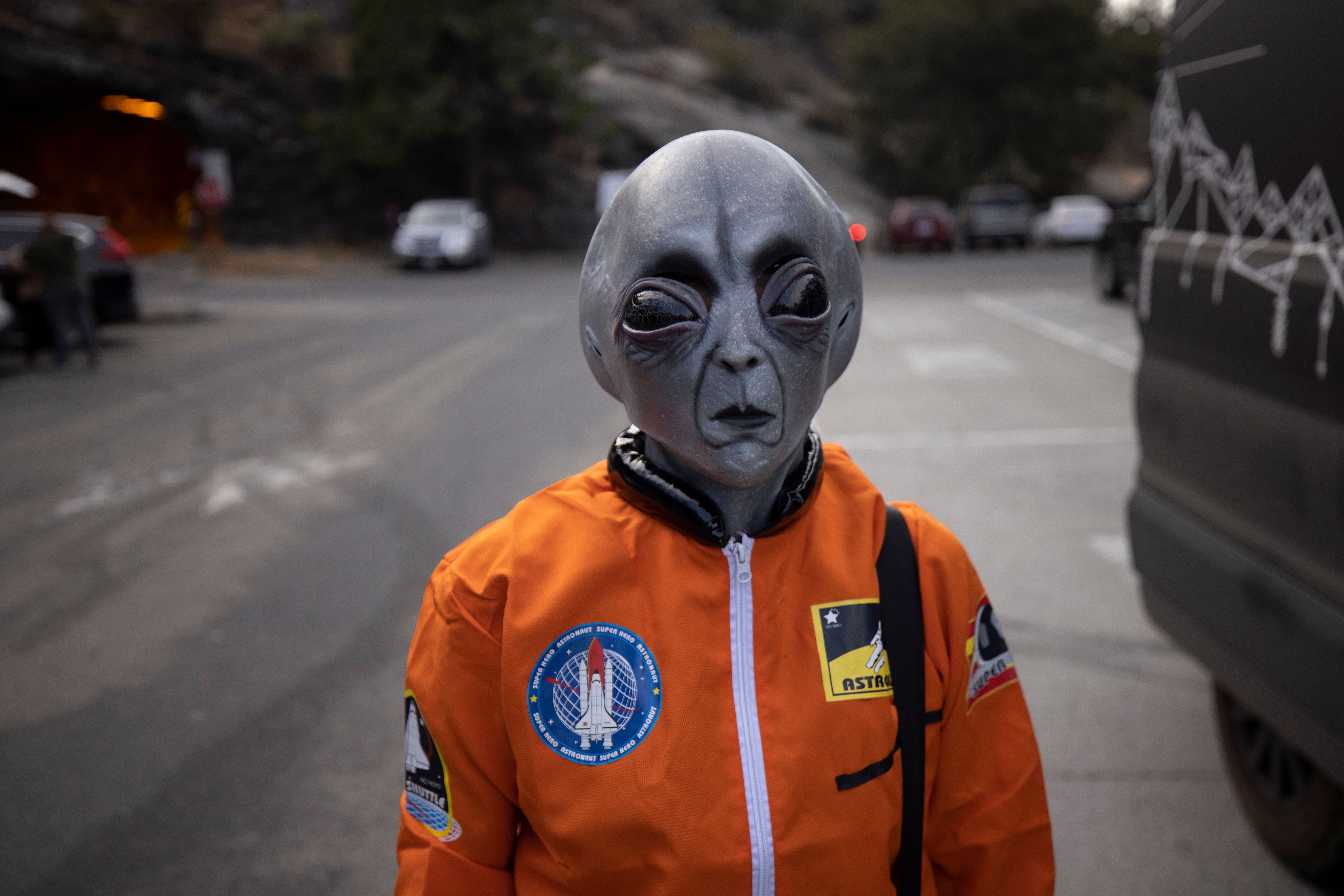 A person in an alien mask and orange astronaut suit stands on a city street during the day.