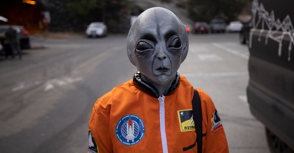 A person in an alien mask and orange astronaut suit stands on a city street during the day.