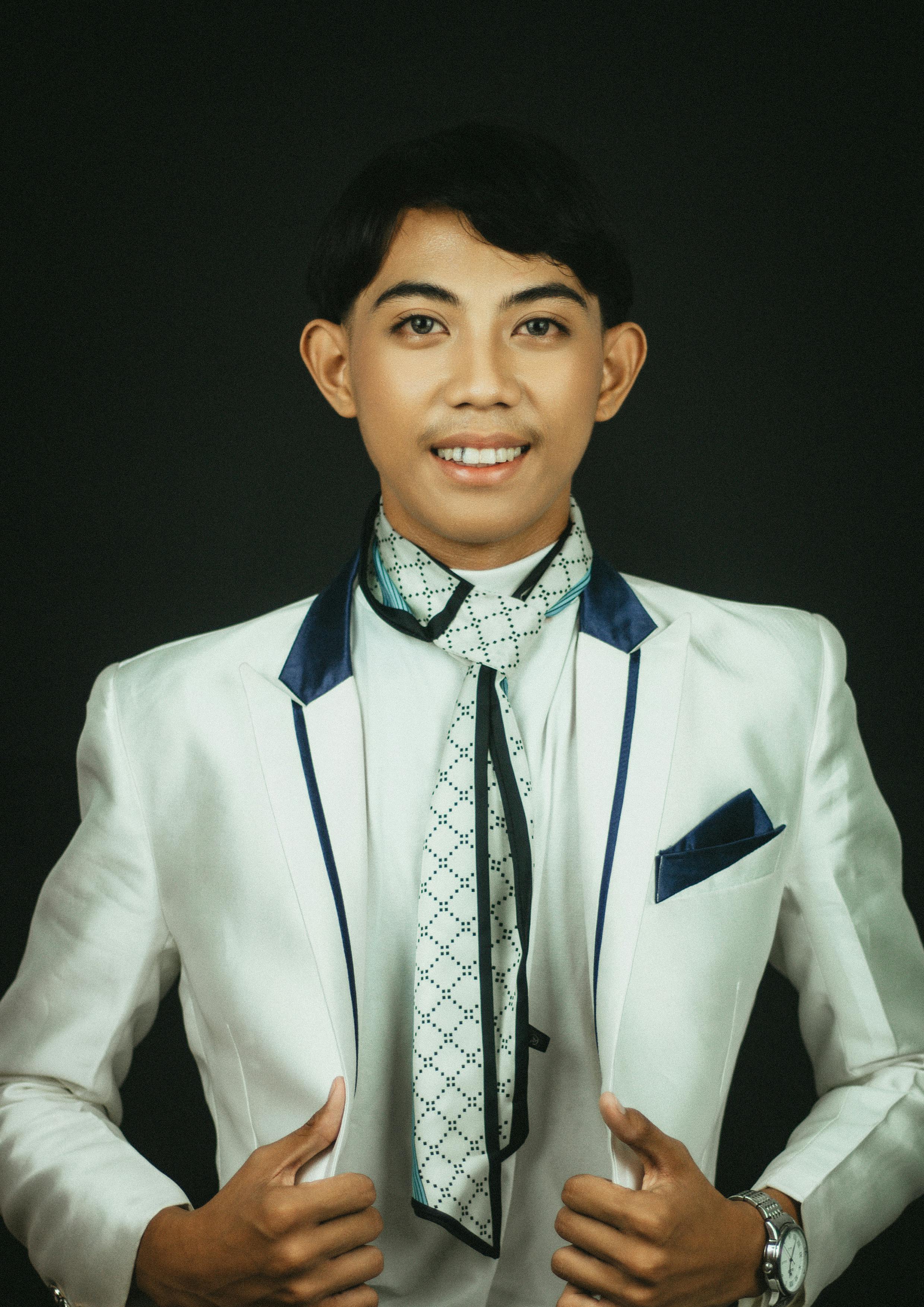 Confident young man dressed in a white formal suit with a unique necktie style, posing against a dark backdrop.