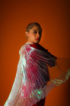 Stylish portrait of a woman draped in a pleated transparent cape against a warm orange background.