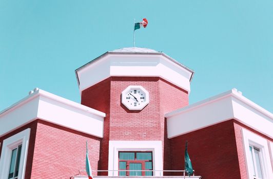 Red brick building with clock tower and flags on a bright day.
