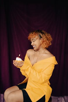 Smiling young woman holding a cupcake with a lit candle against a purple background, celebrating a birthday.