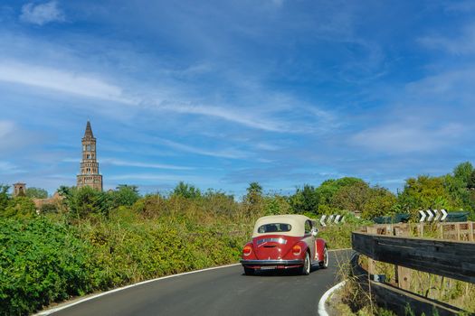 Red vintage car on a countryside road with a historic tower in the background.