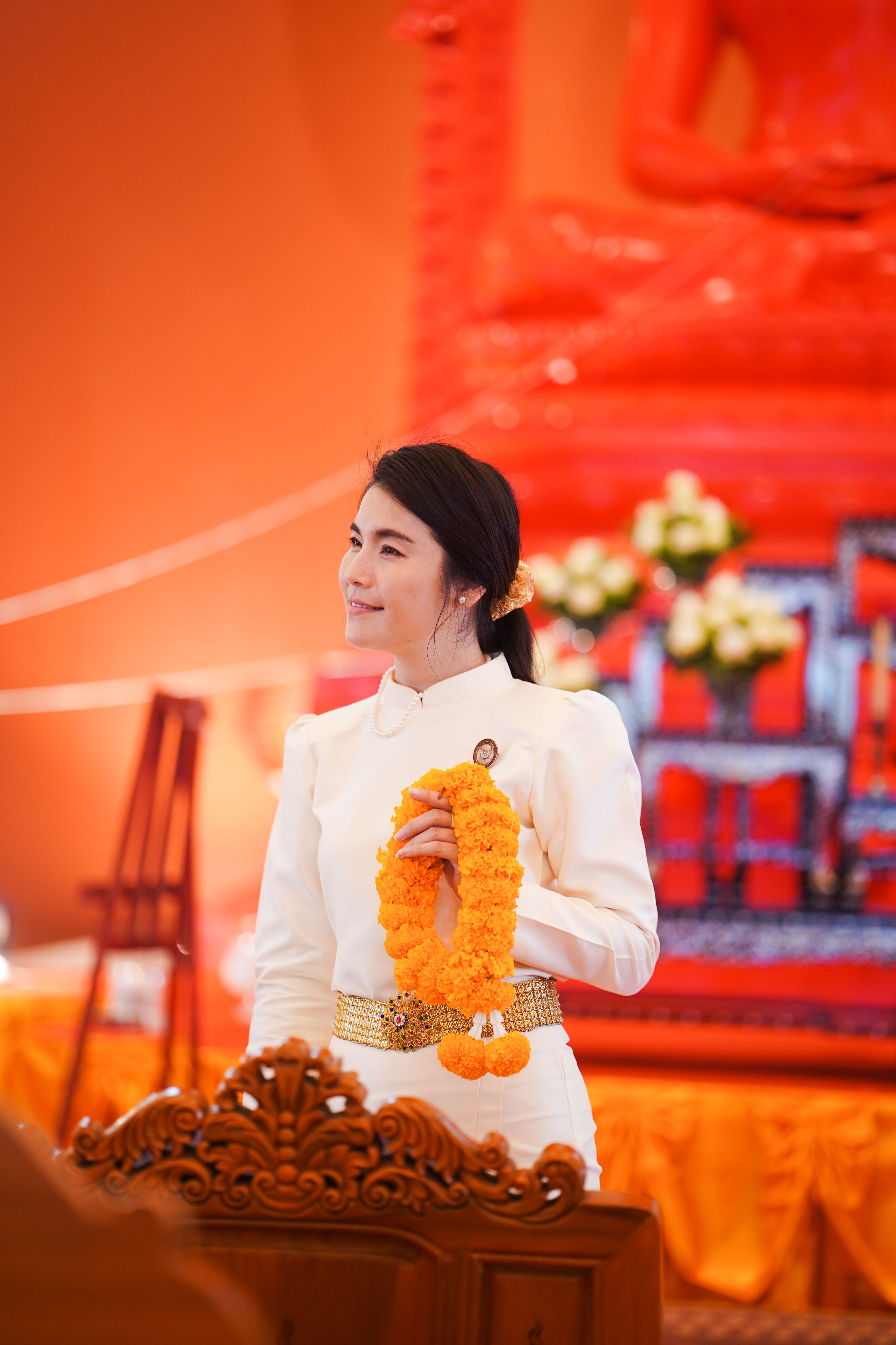 A woman in traditional attire holds a marigold garland during a ceremony in a Thai temple.