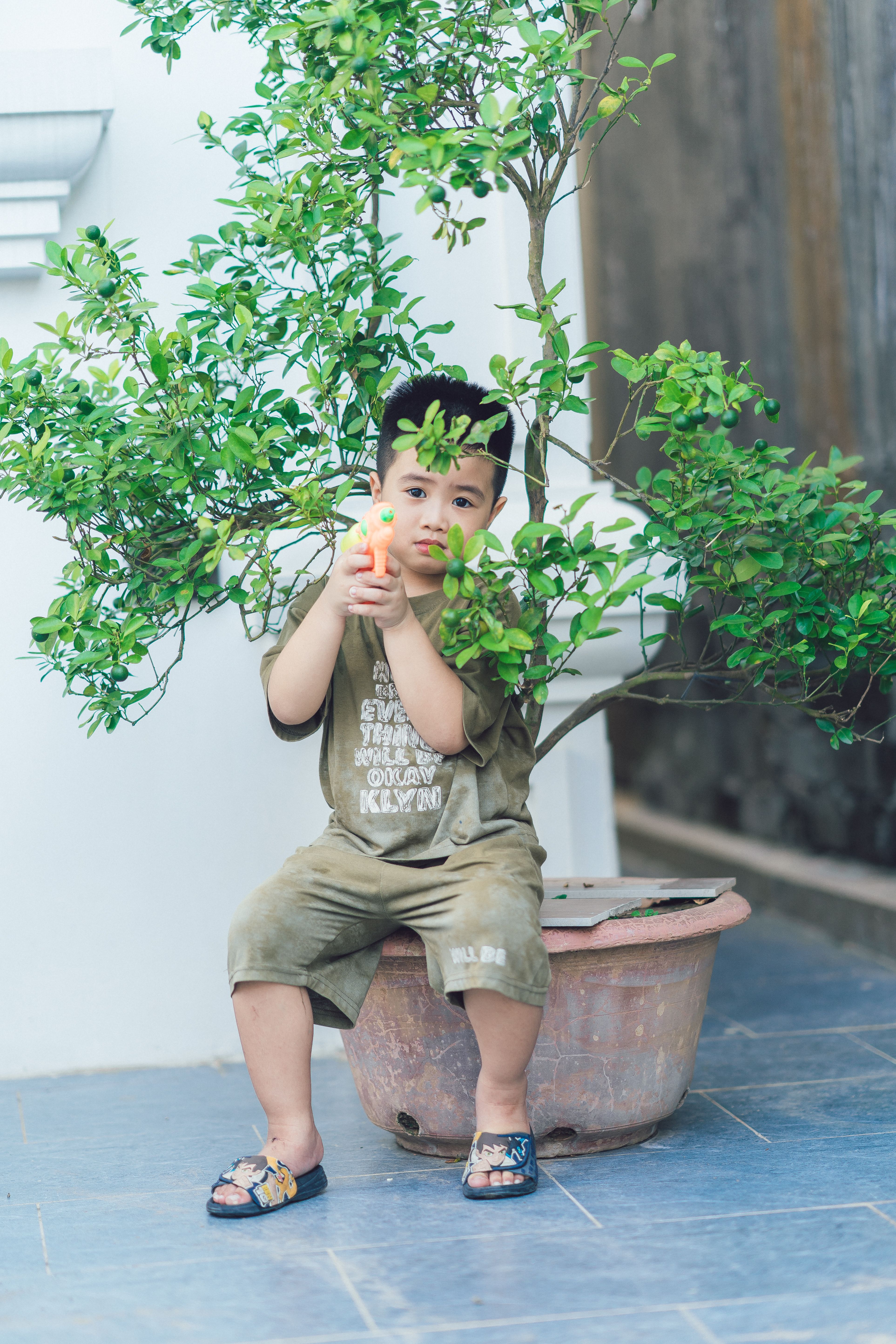Young boy with water gun sitting by potted plant; a playful and candid moment.