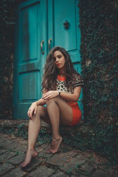 A beautiful woman in stylish attire posing by a teal door with ivy.