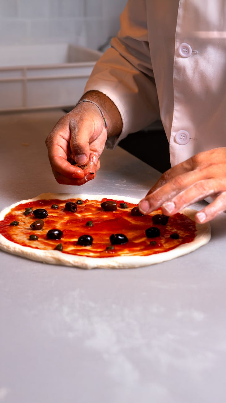 Close-up of a chef adding toppings to fresh pizza dough in a kitchen.