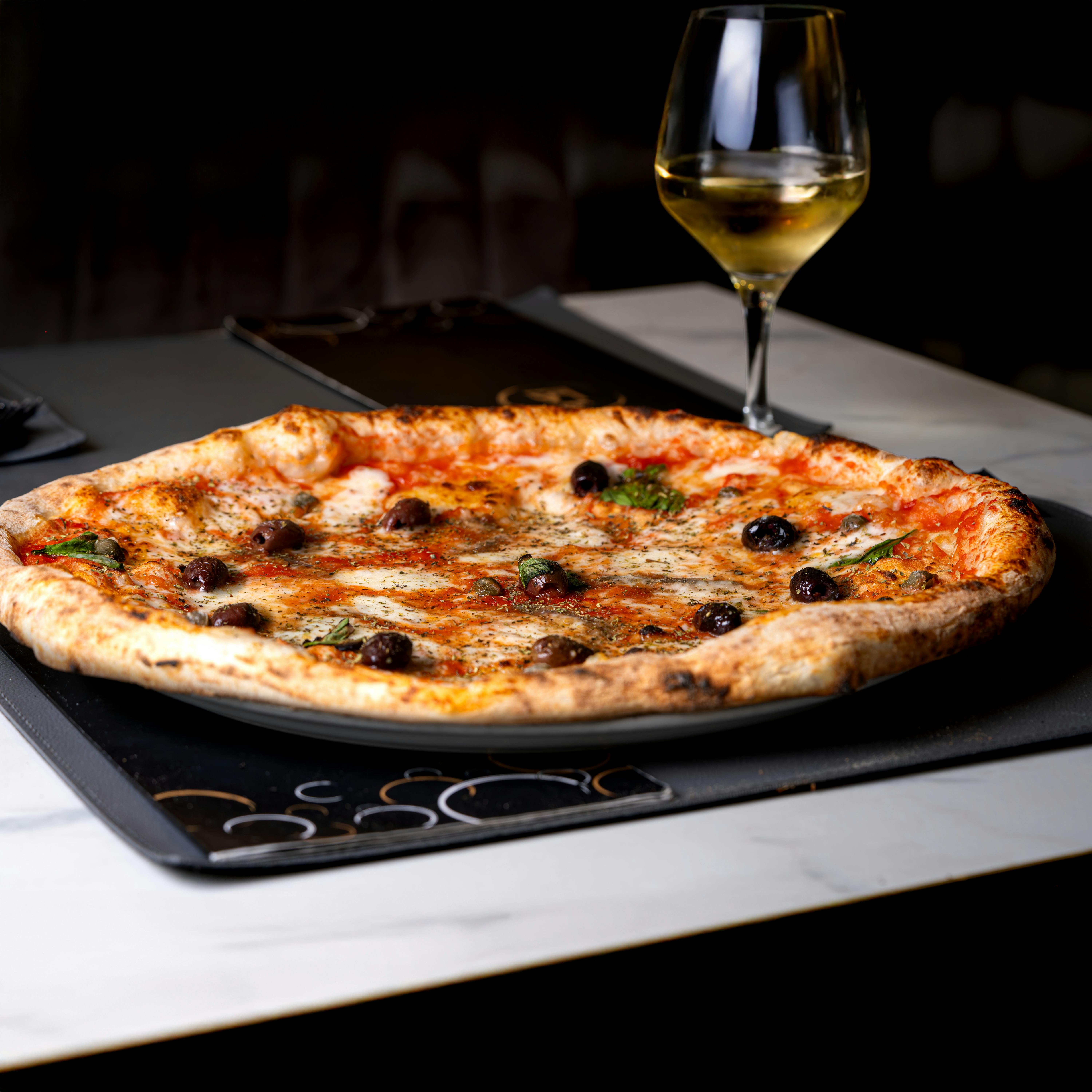 Close-up of a delicious pizza topped with olives and a glass of white wine on a marble table.