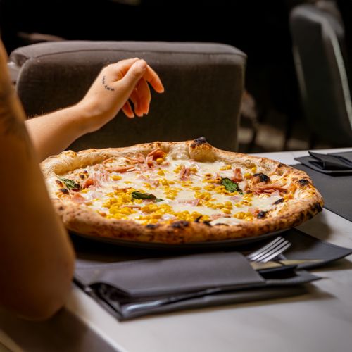 Free Close-up of a ham and corn pizza served in a cozy restaurant with a person reaching for a slice. Stock Photo