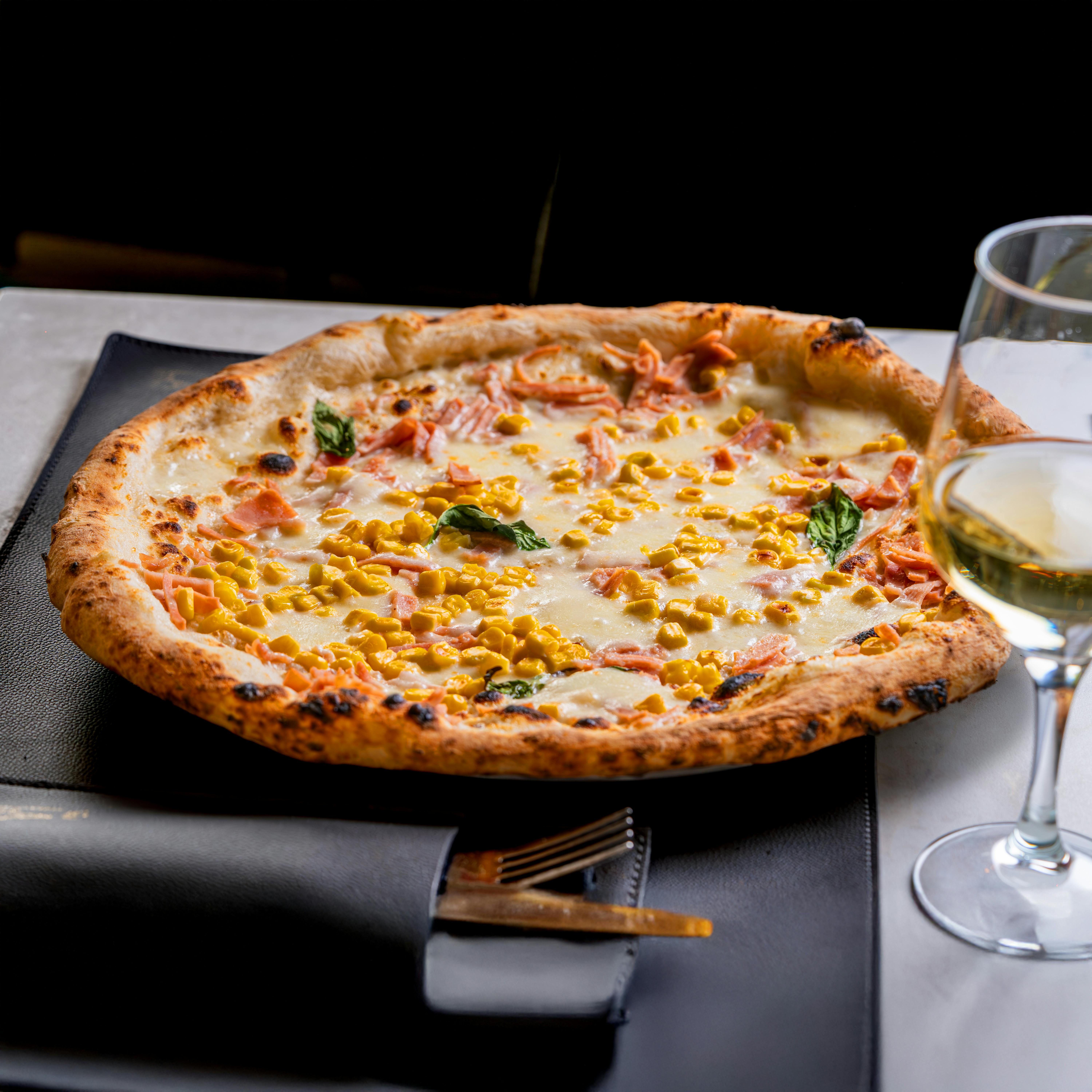 Authentic Italian pizza topped with corn, basil, and cheese, served with a glass of wine.