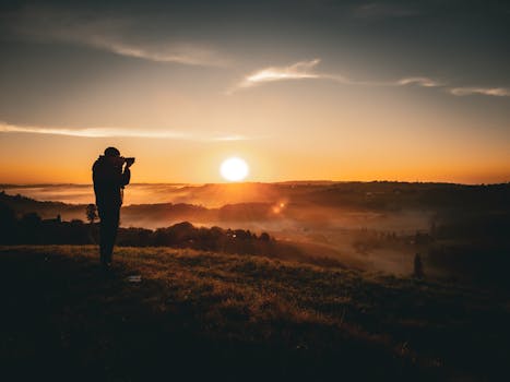 Silhouette of a photographer capturing a tranquil landscape at sunrise. Radiant sun illuminates misty hills in Europe.