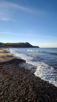 Captivating view of a pebble beach with gentle waves and a distant cliff at sunrise.