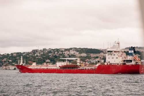 Free A vibrant red cargo ship travels through calm waters with a scenic coastal town in the background. Stock Photo