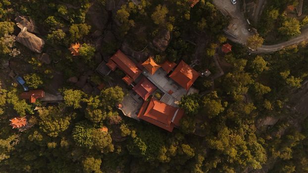 Aerial shot of a temple with red roofs surrounded by dense forest on a hilltop.