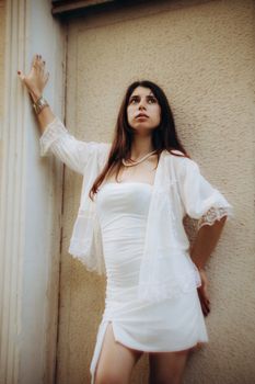 Young woman in a white dress poses elegantly against a wall, exuding grace and sophistication.