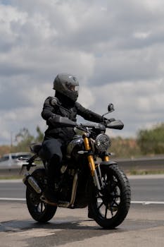 A motorcyclist in black gear rides a motorcycle on an open road under cloudy skies.