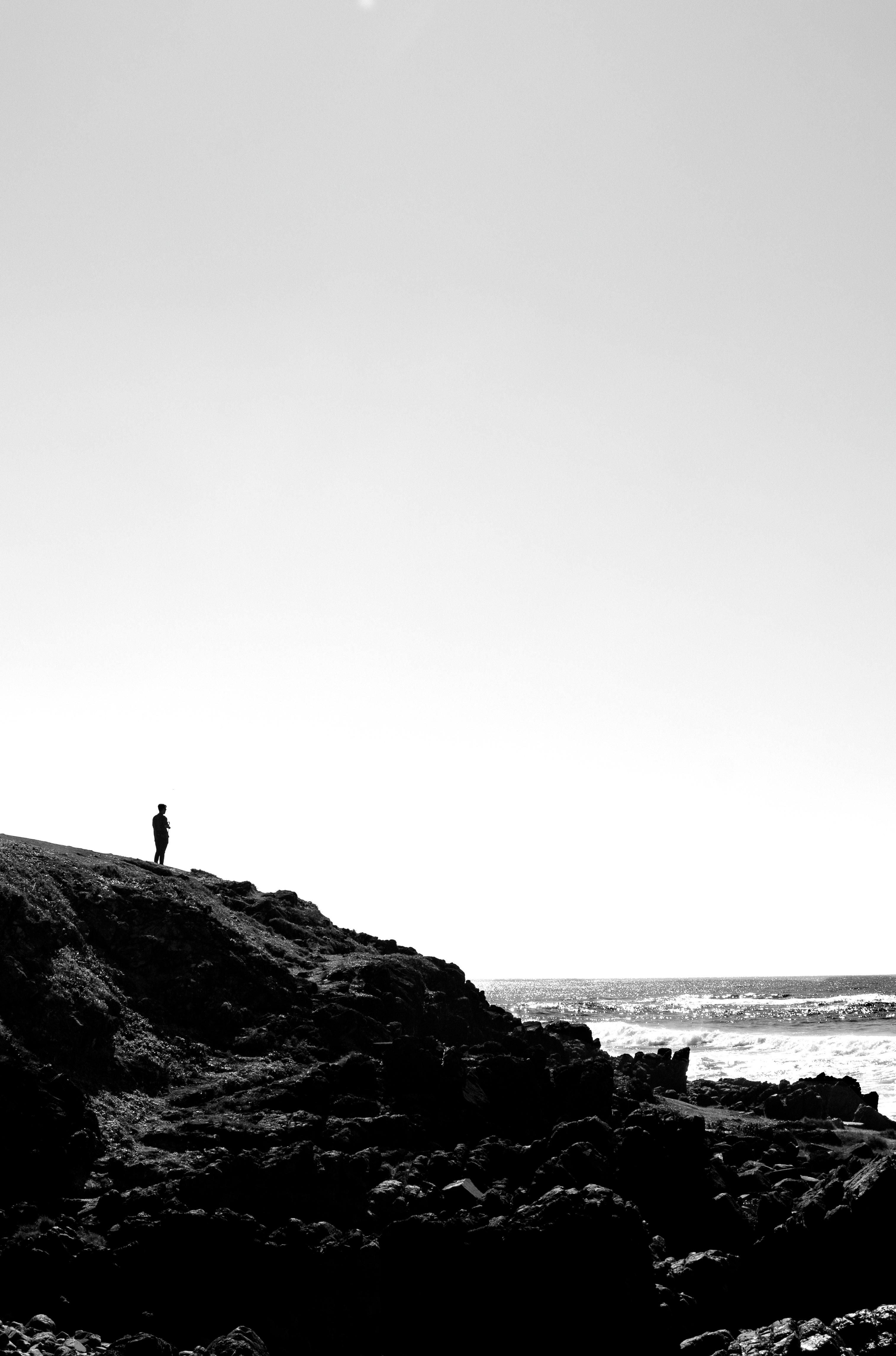 A lone figure stands on a rocky cliff by the ocean, black and white tones.