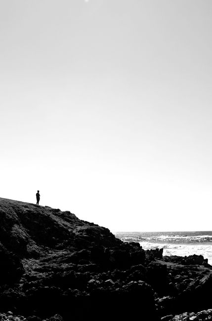 A lone figure stands on a rocky cliff by the ocean, black and white tones.