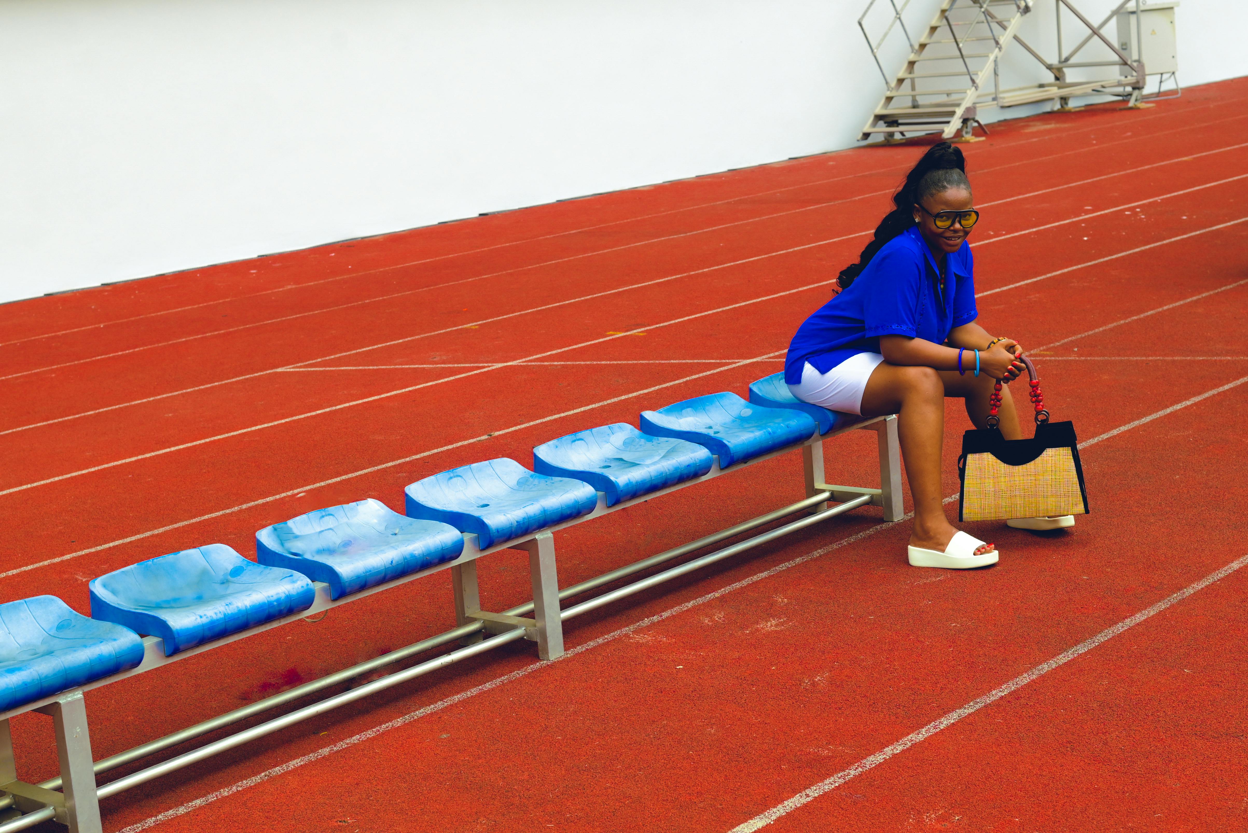 Woman sitting on blue bench in sports track area holding a fashionable handbag.