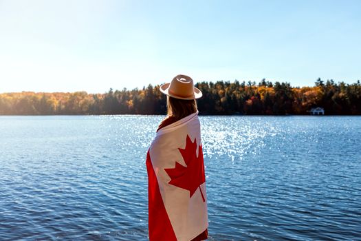 A woman wrapped in a Canadian flag stands by a scenic Ontario lake during fall.