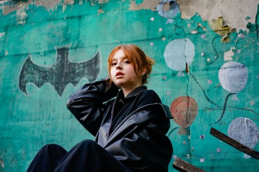 Young woman in black jacket posing before a colorful graffiti wall with bat symbol.