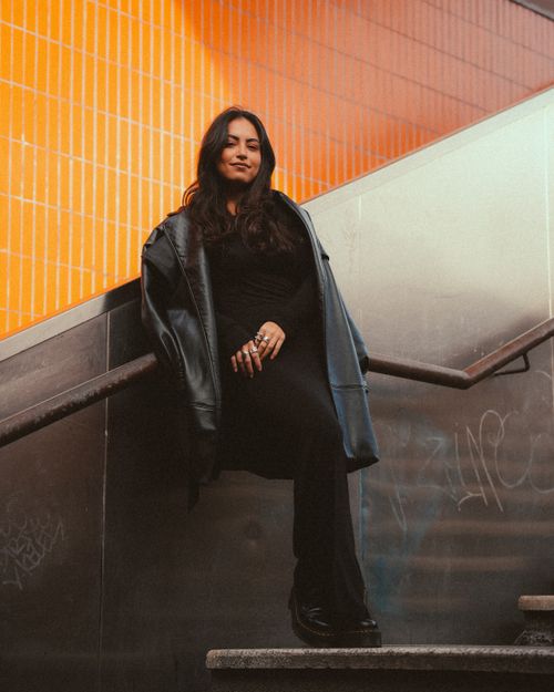 Free Confident woman in a black outfit sits on urban stairs with vibrant orange tile background. Stock Photo