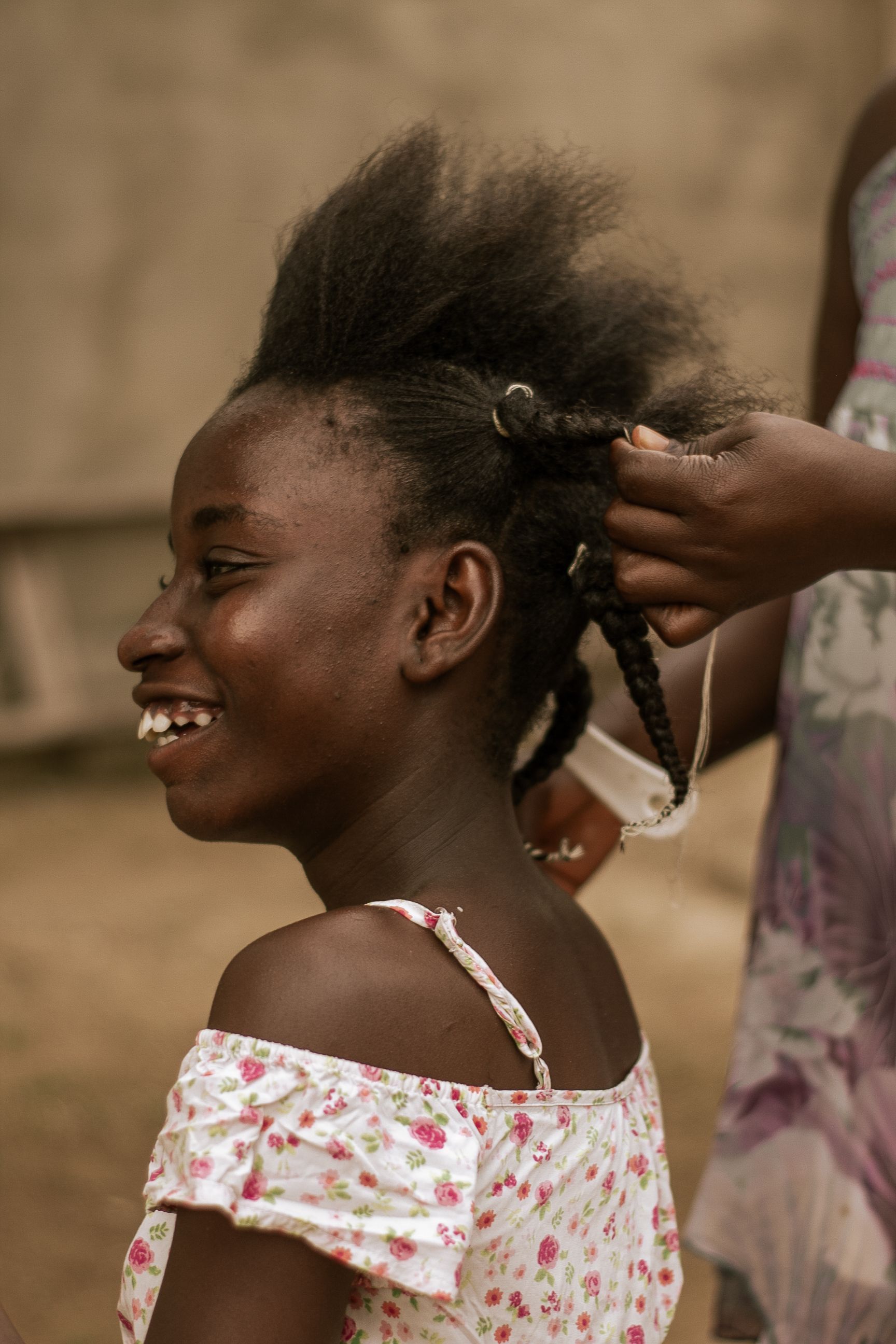 Young girl getting her hair braided outdoors, smiling happily.