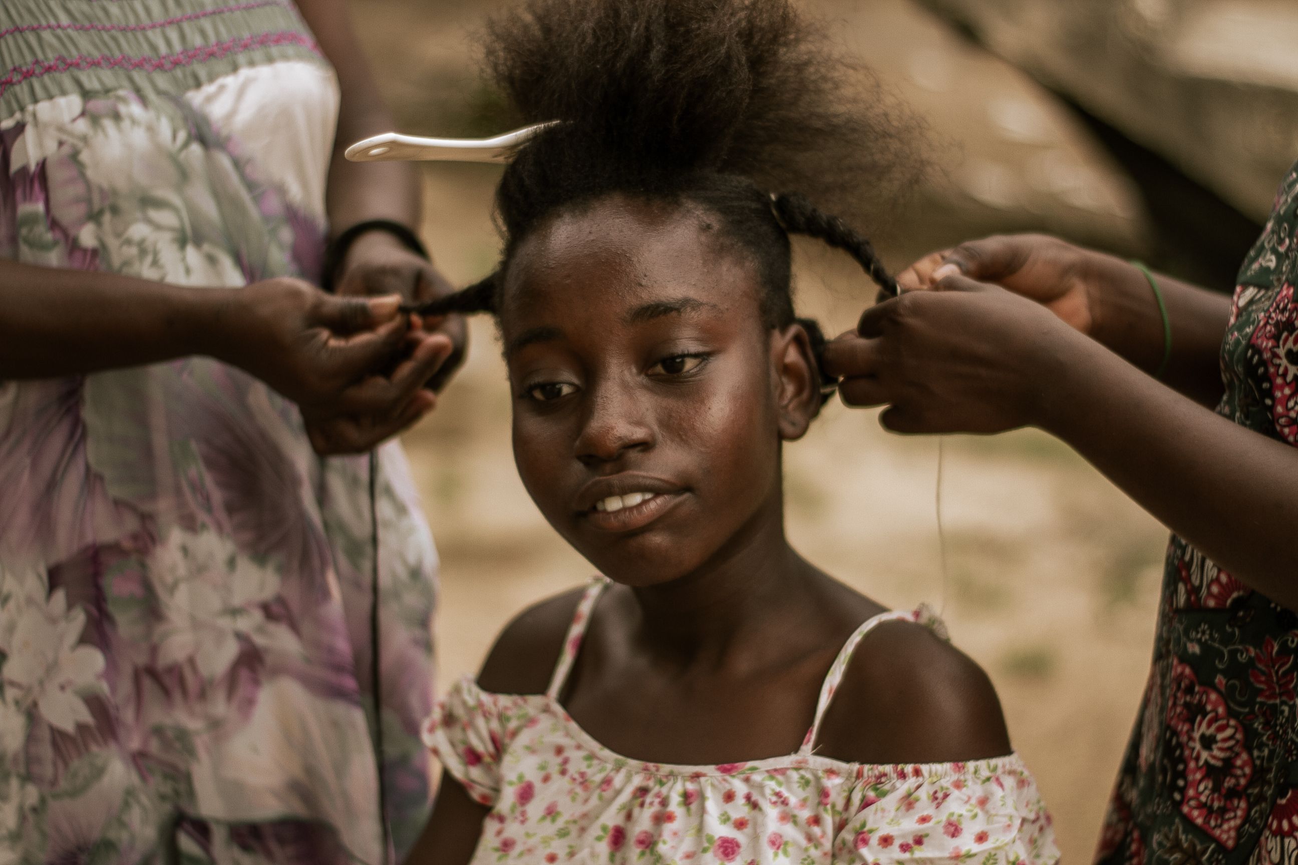 A young girl getting her hair braided by two women in an outdoor setting.