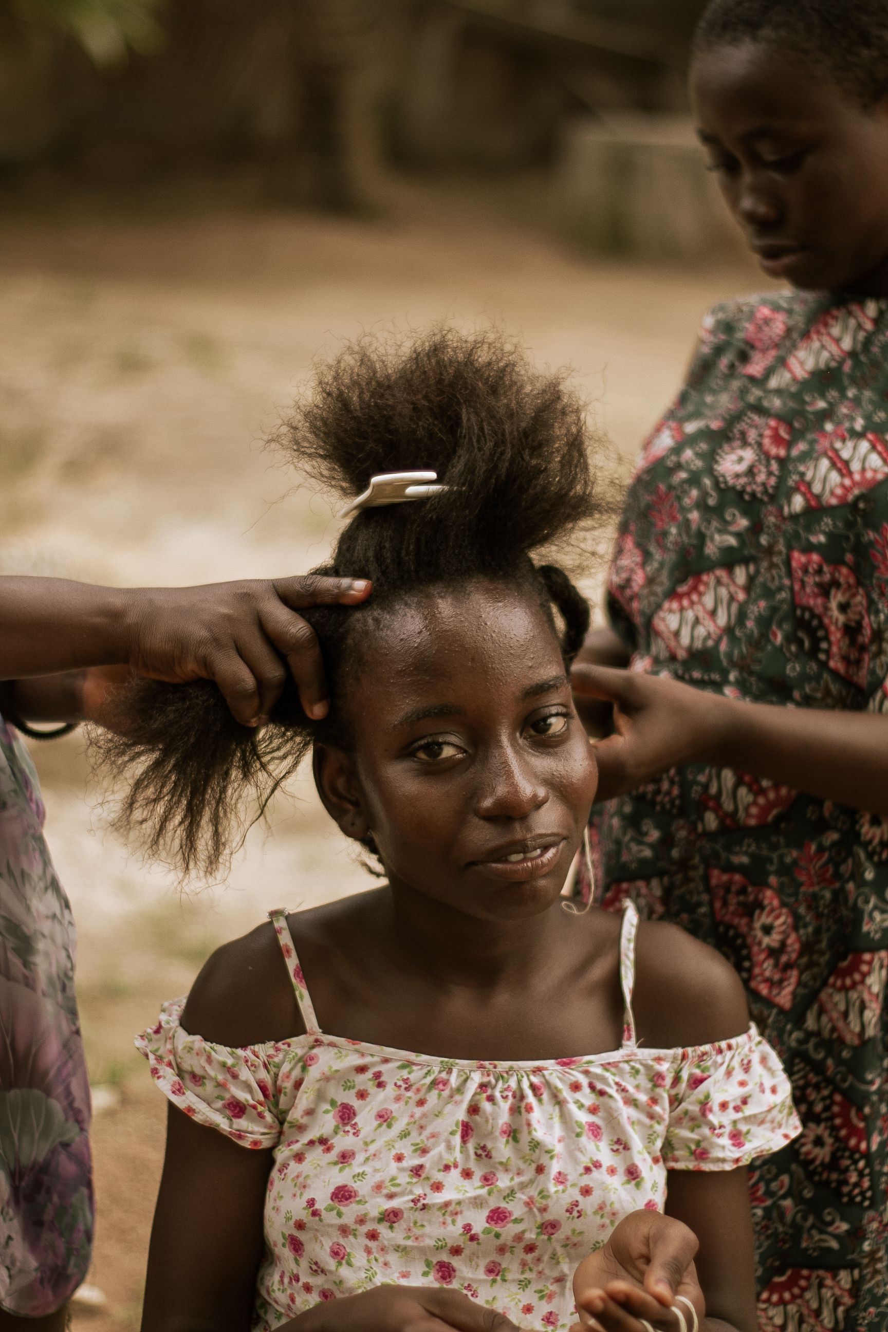 Young girl getting her hair braided outdoors, surrounded by women
