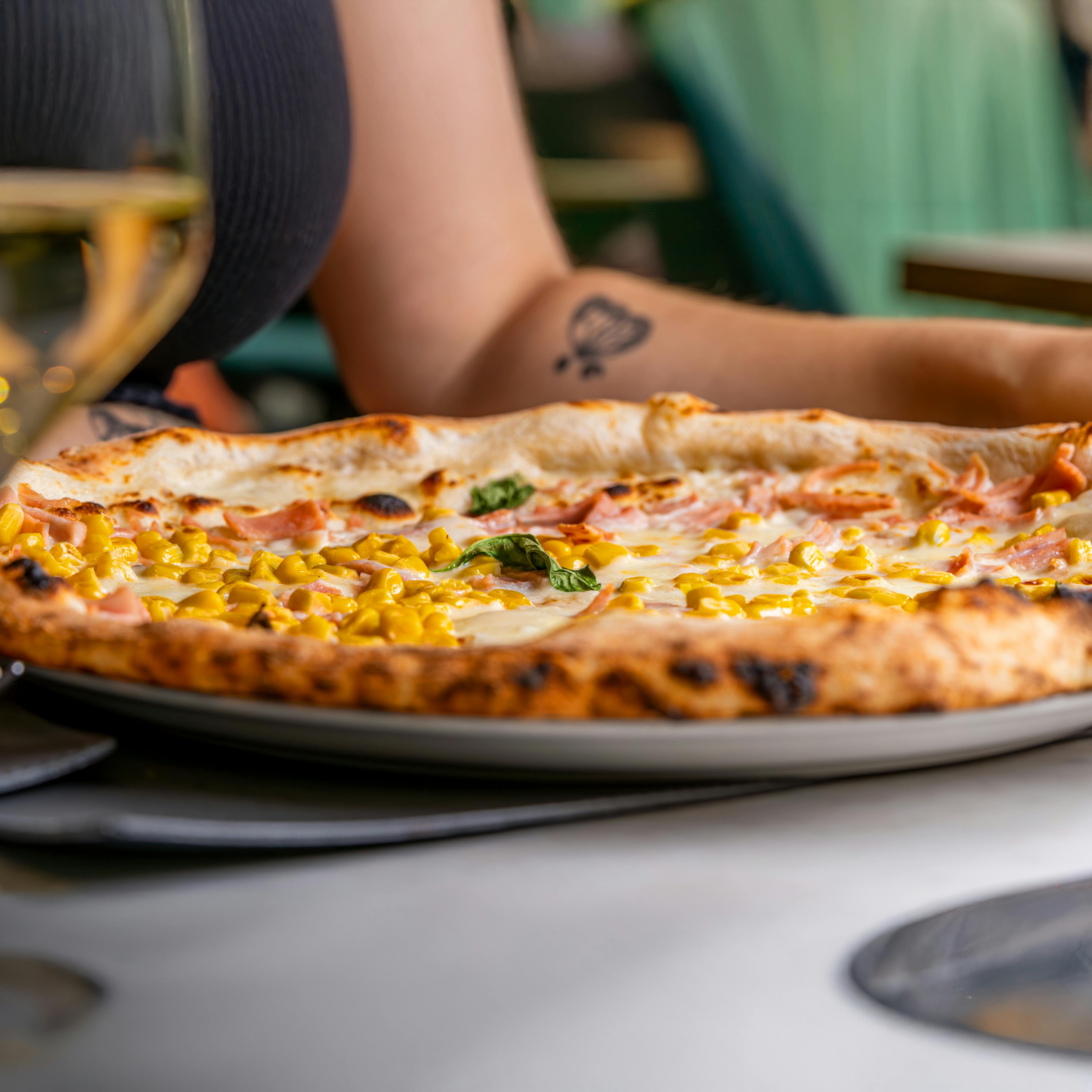 Cheesy corn and ham pizza in a cozy dining setting with tattooed arm.