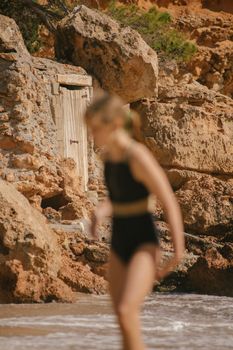 A blurred woman in a swimsuit walks by rocky cliffs at an Ibiza beach.