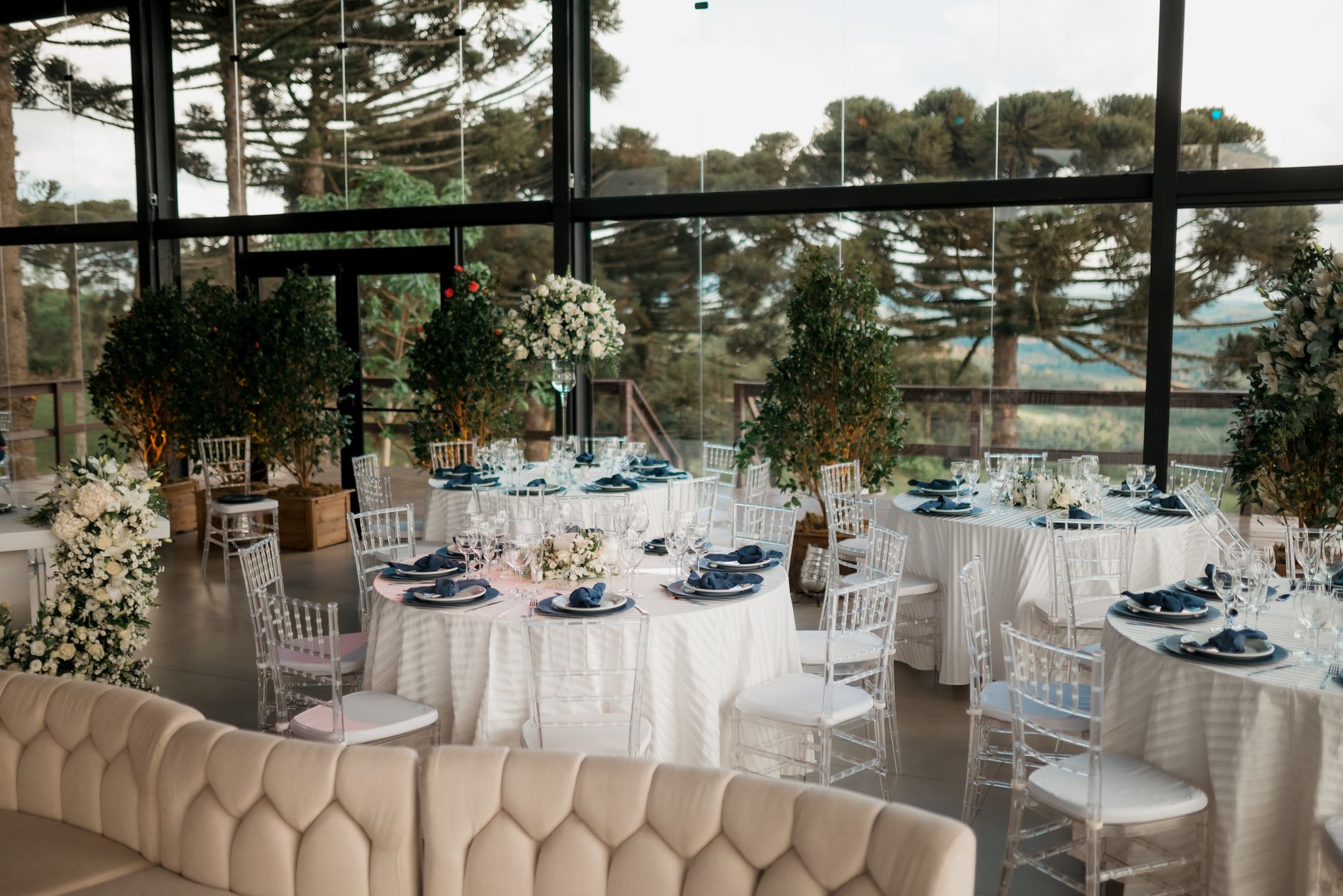 Luxurious wedding reception setup with white tablecloths and floral decorations in a sunlit room with outdoor views.