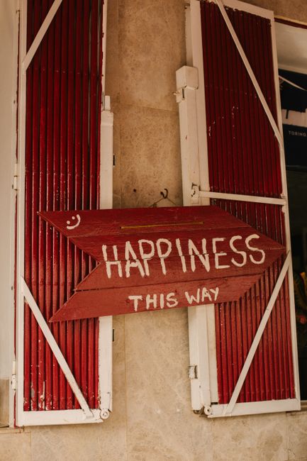 A rustic red arrow sign saying 'Happiness This Way' on a decorative wall with shutters in Ibiza, Spain.