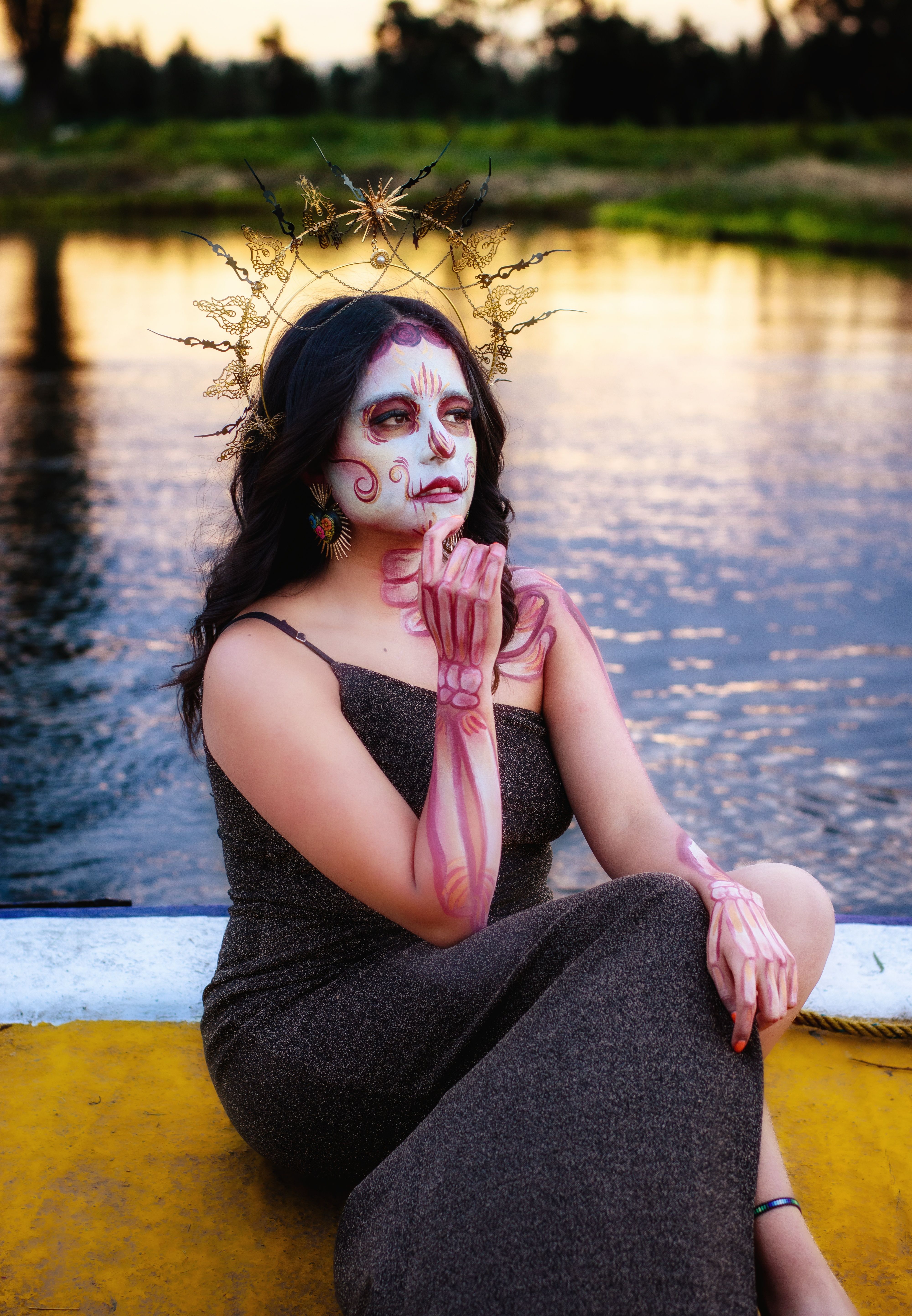 A woman with Dia de Muertos makeup poses elegantly by water during the golden hour.