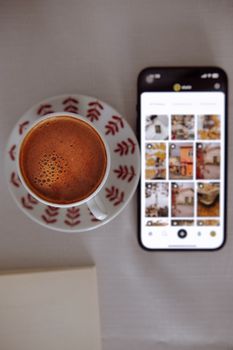 A top view of a cup of coffee beside a smartphone displaying a photo gallery.
