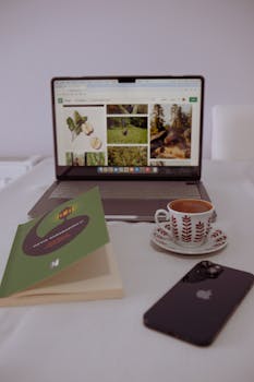 A laptop with a coffee cup beside it, book and phone on a white table.