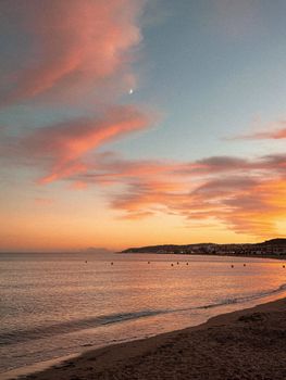 Beautiful beach sunset in Spain with pink clouds, calm sea, and crescent moon.