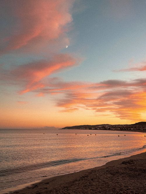 Free Beautiful beach sunset in Spain with pink clouds, calm sea, and crescent moon. Stock Photo