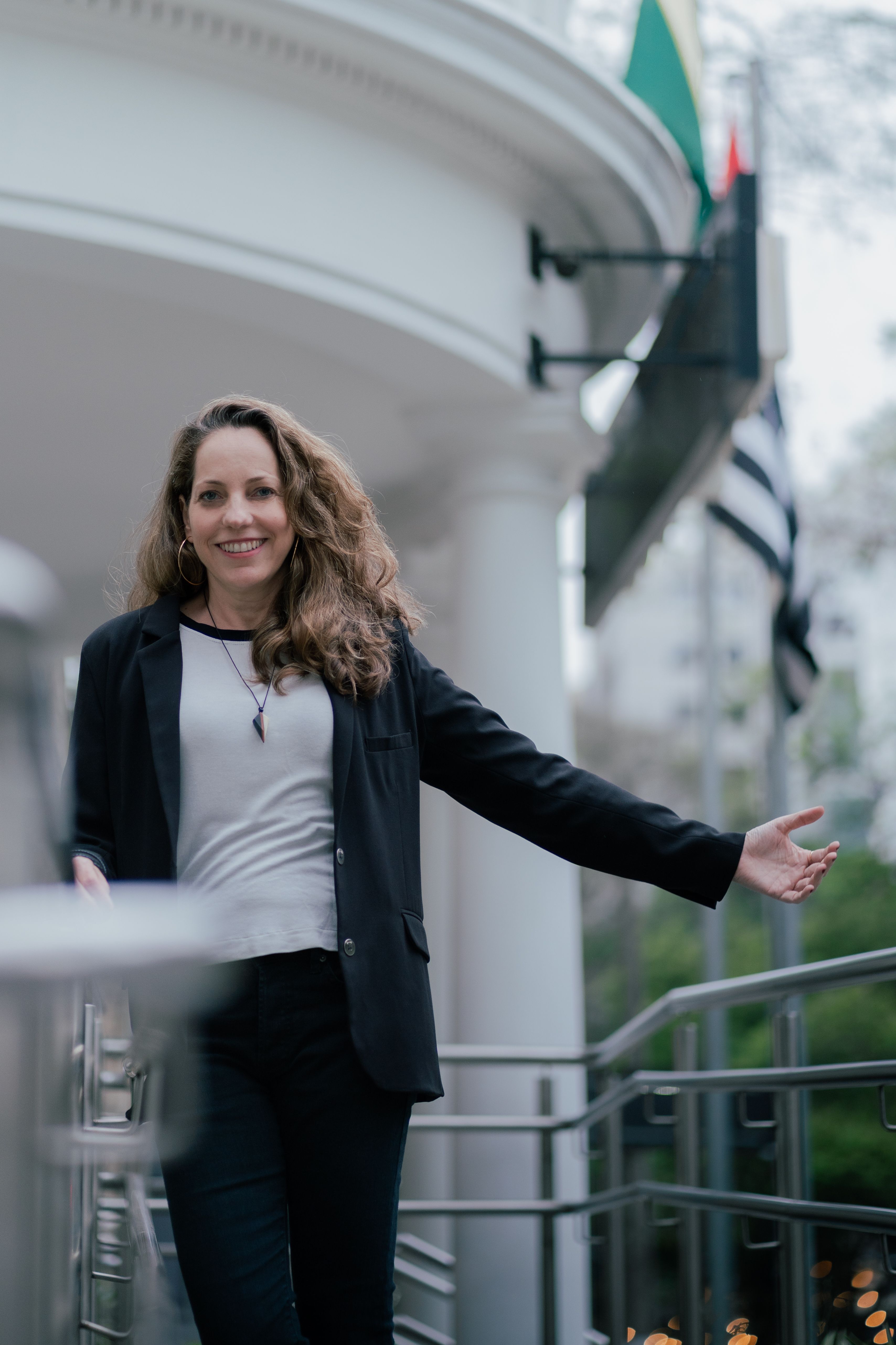 Confident woman in business attire smiling outdoors on a railing with flags in the background.