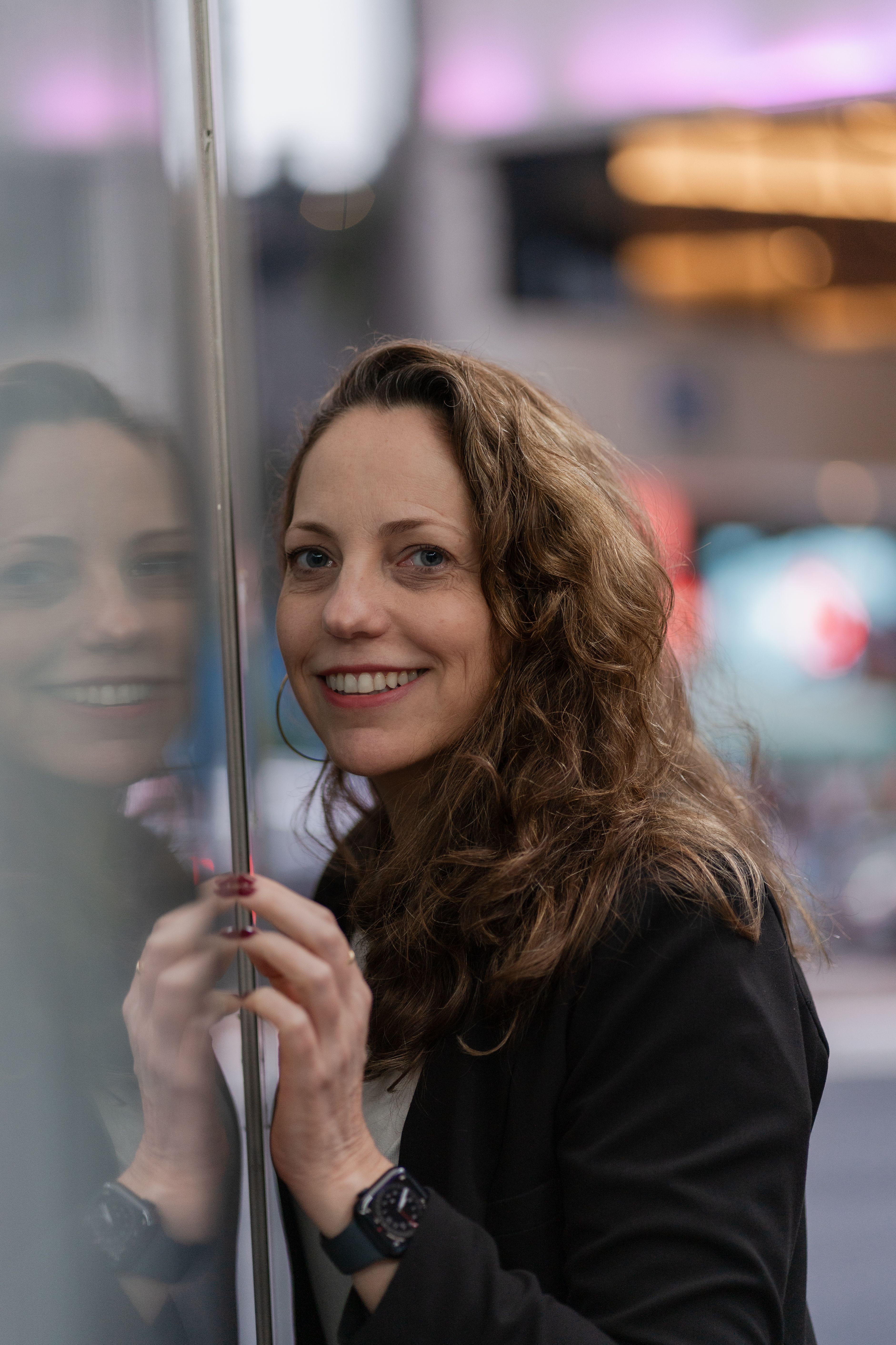 Portrait of a smiling woman with curly hair in a city setting, reflecting on glass.