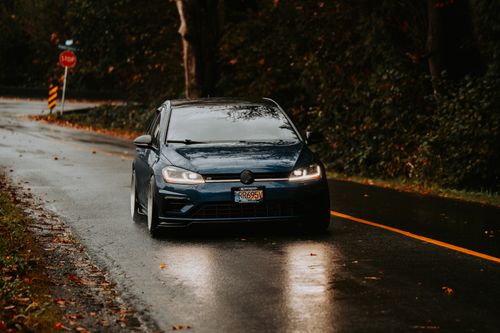 Free Blue car driving on a wet road on a rainy autumn day, surrounded by trees. Stock Photo