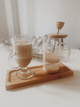 Creamy macchiato served with a glass milk frother on a wooden tray, indoors.