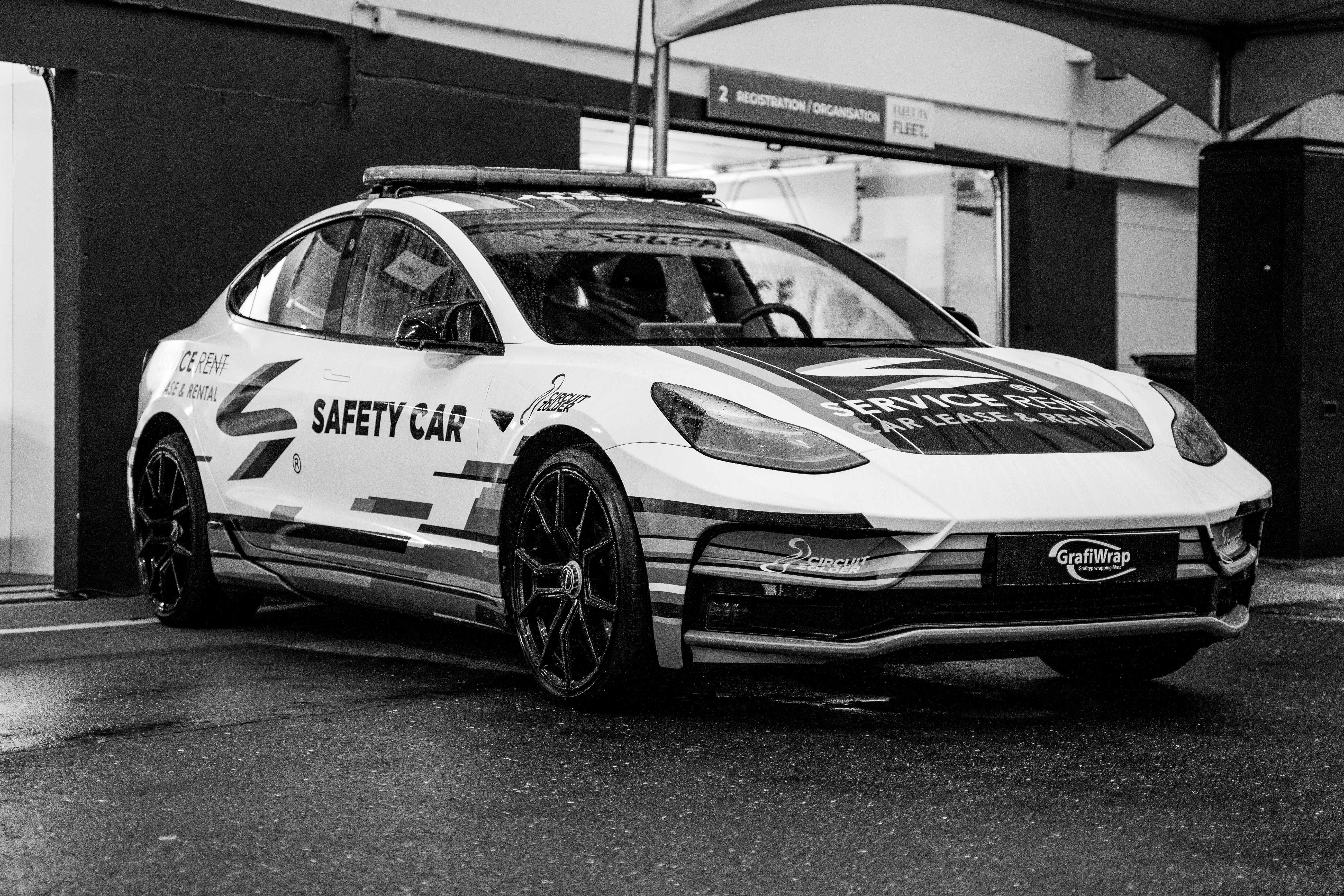 A black and white photo of a Tesla Model 3 safety car parked on a wet track at a racing pit lane.