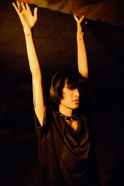 Moody portrait of a young adult with headphones, arms raised, in an underground space.