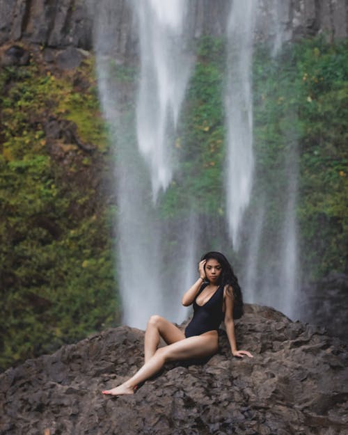Photo of Woman in Black Swimsuit Posing on Rock with Waterfall in the Background